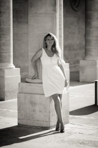 A woman in a white dress leans against a large stone column at Jardin du Palais-Royal, with sunlight casting shadows. The Noir & Blanc image creates a timeless, elegant atmosphere.