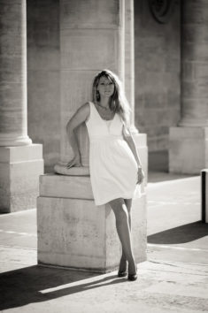 A woman in a white dress leans against a large stone column at Jardin du Palais-Royal, with sunlight casting shadows. The Noir & Blanc image creates a timeless, elegant atmosphere.