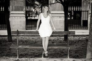 Aurore, in a white dress, sits on the backrest of a wooden park bench, smiling at the camera. Fallen leaves blanket the ground in this Noir & Blanc photo taken at Jardin du Palais-Royal, with trees and an iron fence in the background.