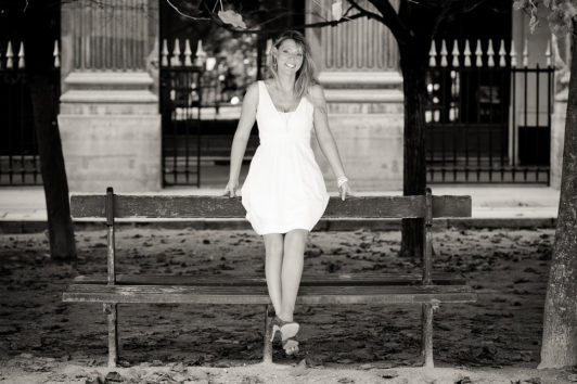 Aurore, in a white dress, sits on the backrest of a wooden park bench, smiling at the camera. Fallen leaves blanket the ground in this Noir & Blanc photo taken at Jardin du Palais-Royal, with trees and an iron fence in the background.