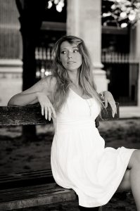 A woman in a white dress sits thoughtfully on a wooden bench outdoors in the Jardin du Palais-Royal. The Noir & Blanc image features blurred columns, trees, and a fence in the background.