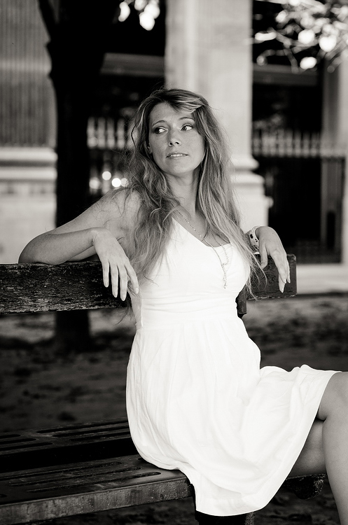 A woman in a white dress sits thoughtfully on a wooden bench outdoors in the Jardin du Palais-Royal. The Noir & Blanc image features blurred columns, trees, and a fence in the background.