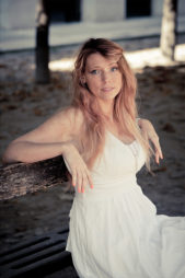 Aurore, in a white sleeveless dress, sits on a park bench at Jardin du Palais-Royal. Her long, wavy light brown hair frames her face as she looks at the camera, with vibrant couleurs from trees and scattered leaves behind her.