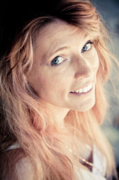 A woman with long, wavy reddish-blonde hair and blue eyes smiles warmly at the camera in the Jardin du Palais-Royal. She is wearing a white top and is lit softly, adding cheerful Couleurs to the bright scene.