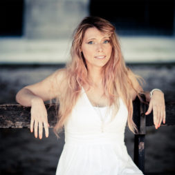 A woman with long, wavy light brown hair sits on a wooden bench at the Jardin du Palais-Royal, wearing a sleeveless white dress and a necklace, looking at the camera with a gentle expression.