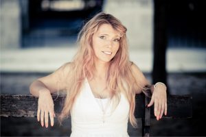 A woman with long, wavy blonde hair wearing a white dress sits on a wooden bench in the Jardin du Palais-Royal, smiling softly at the camera as gentle couleurs play across the scene.