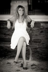A woman with long hair wearing a white dress and heels sits on a wooden bench in the Jardin du Palais-Royal, smiling at the camera. The image captures her elegance in classic Noir & Blanc style.