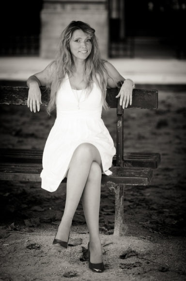 A woman with long hair wearing a white dress and heels sits on a wooden bench in the Jardin du Palais-Royal, smiling at the camera. The image captures her elegance in classic Noir & Blanc style.