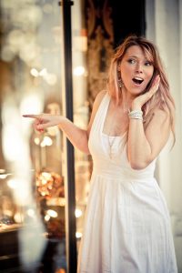 A woman in a white dress stands by a shop window near the Jardin du Palais-Royal, looking surprised and delighted while pointing inside. Her mouth is open in excitement, bracelets and earrings glinting amid softly blurred Couleurs in the background.