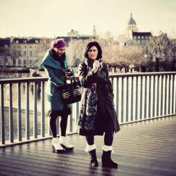 Camilla and Nikki stand on a bridge in the city. Camilla, with short purple hair, wears a teal coat and white boots, holding a handbag. Nikki, with long dark hair, sports a long dark coat, patterned scarf, and black boots. A dome and cityscape are visible in the background.