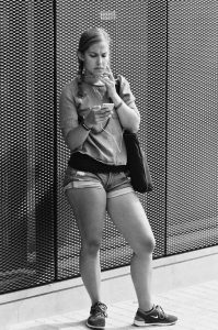 A woman, Helena Gonzales, wearing shorts and a short-sleeve shirt stands against a perforated metal wall, looking at her phone with a focused expression. She has a bag over her shoulder and one hand touching her face.