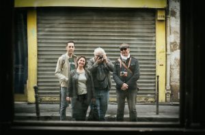 Sebastien, Laurent, and Nathalie stand along with another person in front of a yellow building with a closed, gray shutter, posing for a photo. The person in the middle is taking the picture with a camera. The group appears reflected in a window, suggesting the photo is of their reflection.