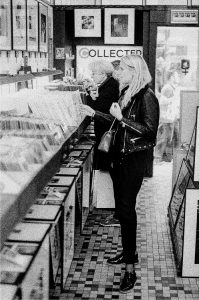 A black-and-white photo of a record store captures the feel of a marché des enfants rouges. A woman with long hair in a leather jacket stands browsing records in the foreground, while an elderly man with glasses and a coat looks through records further down. Framed pictures adorn the walls, giving it a galerie photo vibe.