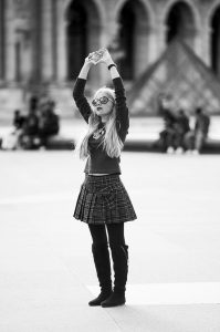 A woman with long hair and sunglasses stands with outstretched arms in a plaza, exuding expression of "mon amour." She is wearing a plaid skirt, black leggings, boots, and a sweater. In the blurred background, people can be seen sitting and walking near an architectural structure.