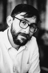 Black and white portrait of a man with short hair, glasses, and a beard. He is wearing a light-colored button-up shirt and is looking at the camera with a slight smile. Captured during a Grain de photographe photo workshop, the background is artistically blurred.
