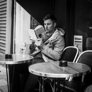 A man with short hair, wearing a hooded jacket, is sitting alone at a small café table outdoors, engrossed in reading Maya Angelou. An empty wine glass sits on the table beside him, and two additional empty chairs are visible.