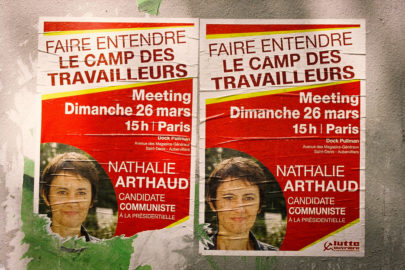 Two posters in French announce a political meeting featuring Nathalie Arthaud, a communist candidate. The event, part of her campaign, is scheduled for Sunday, March 26 at 3 PM in Paris. The posters are affixed to a green, worn surface and include a photo of Arthaud.