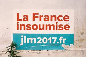 A weathered poster on a wall in Paris reads "La France insoumise" in bold red letters above a blue section containing the text "jlm2017.fr." The bottom right corner of the poster is tattered, and a small plant grows near the lower left edge of the campaign poster.