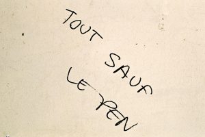 The image shows graffiti on a wall in Paris with the text "TOUT SAUF LE PEN," handwritten in black ink. The words are slightly slanted and scattered across the beige, slightly textured wall. The phrase, which translates to "Anything but Le Pen," captures the local sentiment during the campagne.