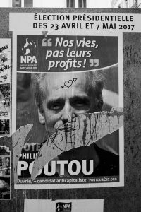 Black and white image of a French election poster for the 2017 presidential campaign in Paris. The poster features Philippe Poutou, with the slogan "Nos vies, pas leurs profits!" (Our lives, not their profits!). The campagne poster is partially torn and defaced with a drawn mustache.