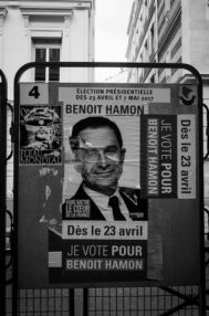 Black and white image of a political campaign poster for Benoît Hamon, promoting his candidacy in the 2017 French presidential election. The poster, seen throughout Paris and the countryside, features his portrait and slogans, including "Je vote pour Benoît Hamon" and "Dès le 23 avril".