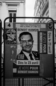 Black and white image of a political campaign poster for Benoît Hamon, promoting his candidacy in the 2017 French presidential election. The poster, seen throughout Paris and the countryside, features his portrait and slogans, including "Je vote pour Benoît Hamon" and "Dès le 23 avril".