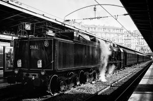 A black and white image of a vintage steam locomotive halted at Gare de l'Est platform in Paris. The Train Spécial Vapeur emits steam, and overhead electric lines are visible. The station has a metal canopy, and buildings can be seen in the background.