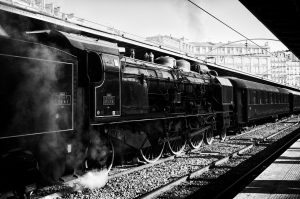 Black and white photo of a vintage steam locomotive at Gare de l'Est in Paris. The Train Spécial Vapeur is stationary on the tracks, with steam emitting from below the engine. The station has a covered platform, and buildings are visible in the background.