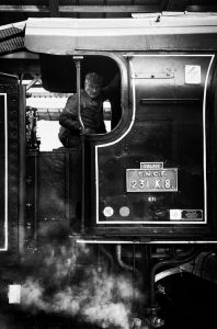 A black and white photograph shows a man standing inside the cab of a vintage steam locomotive at Gare de l'Est in Paris. He holds onto the doorframe with one hand, looking slightly downwards. The train's machinery is prominently visible, with steam billowing below the cab of the Train Spécial Vapeur.
