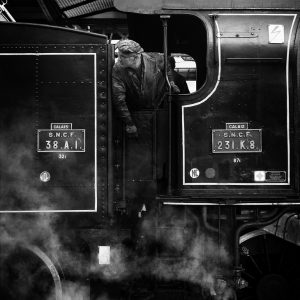 Black and white photo of a person leaning out of a steam locomotive's cab at Gare de l'Est. The person, wearing a cap and work clothes, inspects or looks forward. The locomotive, part of the Train Spécial Vapeur, has signage reading "S.N.C.F." and "Calais." Steam surrounds the lower part of the engine.