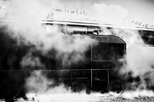 Black and white photo of a Train Spécial Vapeur emitting large clouds of steam. The train's dark body contrasts with the white steam, obscuring much of its detail. Overhead, the silhouette of Gare de l'Est and nearby buildings is visible under a bright Paris sky.