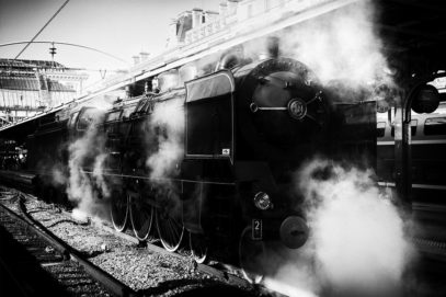 Black and white photo of a steam locomotive at Gare de l'Est in Paris. The Train Spécial Vapeur engine is emitting clouds of steam, creating a dramatic, nostalgic atmosphere. Overhead, a glass roof shelters the platform, and several carriages are visible in the background.