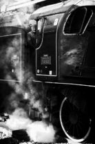 A black and white photo shows a steam locomotive billowing smoke at Paris's Gare de l'Est. The engine number, 32678, is visible as a person wearing glasses and a cap leans out of the Train Spécial Vapeur’s side window, smiling. The large wheels and mechanical details of the train are prominent.