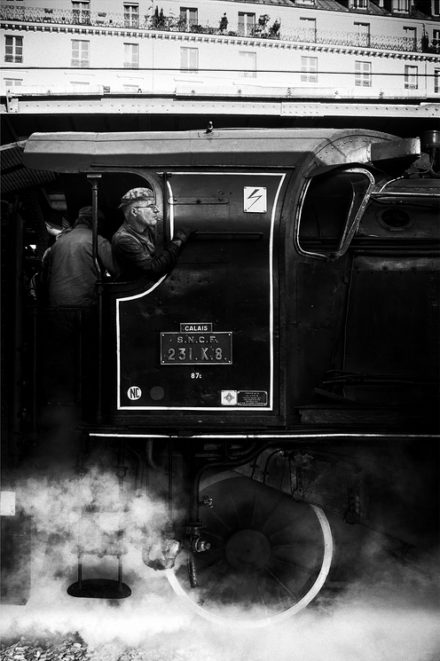A black-and-white photo depicts a steam locomotive with billowing smoke. Two workers are visible, one looking out of the window, the other stands outside. The train is labeled "Calais S.N.C.F 231.K.8". A building with windows and railings is in the background—likely Gare de l'Est in Paris.
