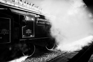 Black and white image of a Train Spécial Vapeur at Paris's Gare de l'est station, emitting clouds of steam. The train's intricate details and wheels are visible, with a person leaning out of the cabin. The station platform and building rooftops are seen in the background.