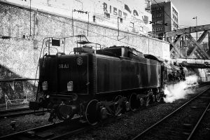 A black-and-white photograph captures a steam locomotive on train tracks at Gare de l'Est in Paris. Steam billows from the engine, set against an urban backdrop featuring tall buildings, a bridge, and a graffiti-covered wall. The number "38A1" is visible on the back of this Train Spécial Vapeur.