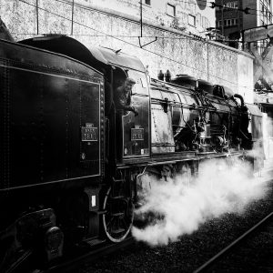 Black and white photo of a Train Spécial Vapeur with a person looking out from the cabin. Steam billows from beneath the locomotive. In the background, classic urban buildings and a bridge overhead evoke the charm of old Paris near Gare de l'Est. The train has visible details and old-fashioned design elements.