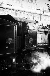 A black-and-white photo featuring two men in the cabin of a vintage locomotive with "S. N. C. F. 231 K 8" written on the side. Steam is visible near the bottom, and graffiti can be seen on the wall above at Gare de l'Est in Paris. Both men are wearing dark clothes and hats, adding to the nostalgic ambiance of this Train Spécial