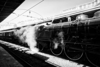 A black and white photo of a Train Spécial Vapeur at Gare de l'Est, emitting steam from its engine. The station platform is partially visible on the left, with overhead wires and the station structure in the background. The scene beautifully captures a vintage atmosphere in Paris.