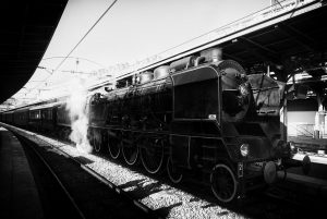 Black and white image of a vintage steam locomotive at Gare de l'Est in Paris. The Train Spécial Vapeur emits a plume of steam and is surrounded by tracks and platforms under a large canopy. The scene is captured from a side angle, highlighting the train’s intricate details.
