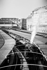 A black-and-white photo of a busy Gare de l'Est train station in Paris with multiple tracks and an old-fashioned Train Spécial Vapeur emitting smoke. People are scattered along the platforms next to the trains, and a large building is visible in the background.