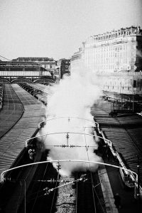 Black and white photograph of the Gare de l'Est in Paris with steam rising from a Train Spécial Vapeur. The station platforms are empty, and there's a row of buildings in the background under a clear sky. The station has an arched roof structure over part of the tracks.