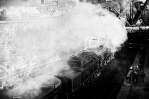 Black and white image of a steam train emitting smoke as it travels along tracks beneath a graffitied wall and metal bridge, reminiscent of the industrial area near Gare de l'Est in Paris. An additional Train Spécial Vapeur is stationed on a separate track to the right, enhancing the historical ambiance.