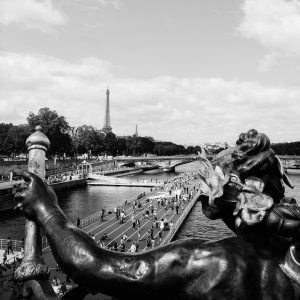 Black and white photo of the Pont Alexandre III bridge in Paris, with a distant view of the Eiffel Tower. The bridge's ornate details and statues are in the foreground, while people walk and cycle along the bridge and riverbank, evoking an Olympic spirit as if carrying a symbolic flame.