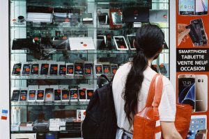 A person with long dark hair wearing a white shirt and carrying a red bag looks at smartphones and tablets displayed in a glass case at the magasin de téléphone. The display includes a variety of devices with red price tags, while an orange sign features promotions.
