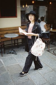 A person in a black hat and outfit walks on a paved street, holding an open magazine or map. They have a white backpack with various decorations, evoking the feel of "Sans Titre." They pass by a café with an empty outdoor table and chairs. The street setting is lined with stone tiles and framed by a building exterior.