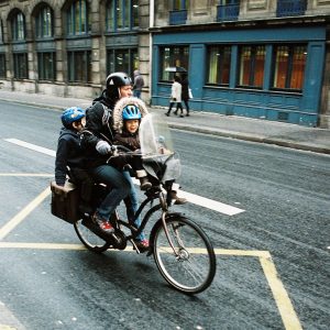 A person riding a vélo with two children seated in front on a busy city street. The person and the children, who appear to be en route with their famille, are wearing helmets. The older child sits behind the handlebars, while the younger one is in front. Buildings and pedestrians blur by as they go vite.