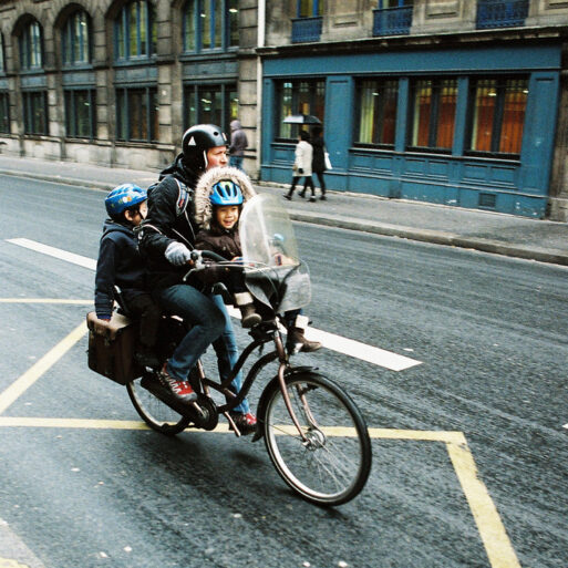 A person riding a vélo with two children seated in front on a busy city street. The person and the children, who appear to be en route with their famille, are wearing helmets. The older child sits behind the handlebars, while the younger one is in front. Buildings and pedestrians blur by as they go vite.