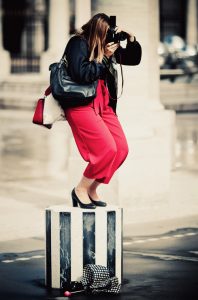 A person with long brown hair is standing on a black-and-white striped pedestal, bent over while taking a photograph with a camera. They are wearing a black jacket, rouge pantalon, and black shoes. On the ground, there is an umbrella with a polka dot pattern.