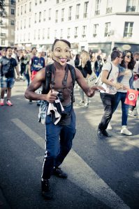 A person wearing a mask and a partially open jacket, revealing their torso, walks towards the camera during the Techno Parade, Edition 2017. People in the background appear to be part of a lively crowd, with buildings and trees visible in the background.